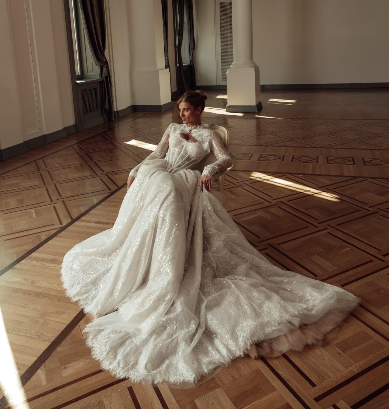 A bride seated in an armchair with a large lace wedding dress whose train spreads across the floor